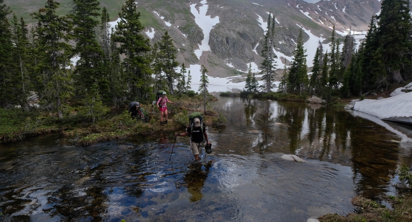 A person wades through shallow water while another person stands on the shore. Behind them are evergreen trees and mountains. 